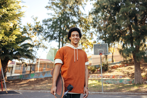 Happy skateboarder carrying his skateboard outdoors during the day. Cheerful teenage boy smiling at the camera while standing outdoors. Sporty teenager wearing casual clothing during the day.