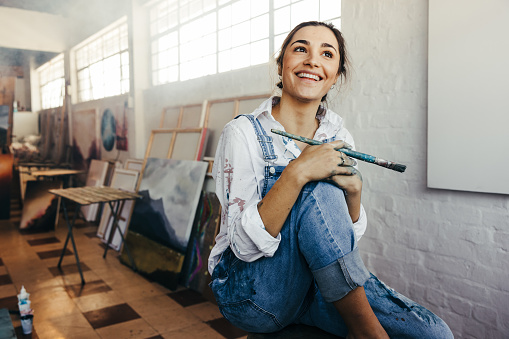 Excited female painter smiling in her art studio. Cheerful young artist holding a paintbrush while sitting in front of a blank canvas. Creative young woman starting a new art project.