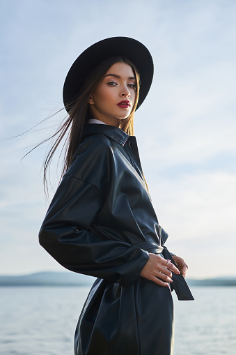 Fashion woman in black round hat and leather raincoat near lake pond. Portrait girl at sunset. Beautiful makeup and bright red lipstick on lips