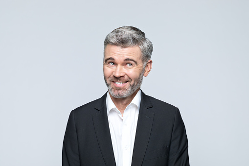 Portrait of handsome mature man wearing black jacket and white shirt, looking up and laughing. Studio shot of male entrepreneur against grey background.