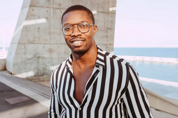 Handsome black man with goatee style looking at camera, wearing eyesglasses, black and white striped shirt, under structure of solar panels in front of the sea. Lifestyle concept.