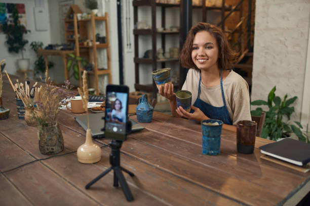 istock Happy young creative female with clay mugs looking at smartphone camera 1353393604