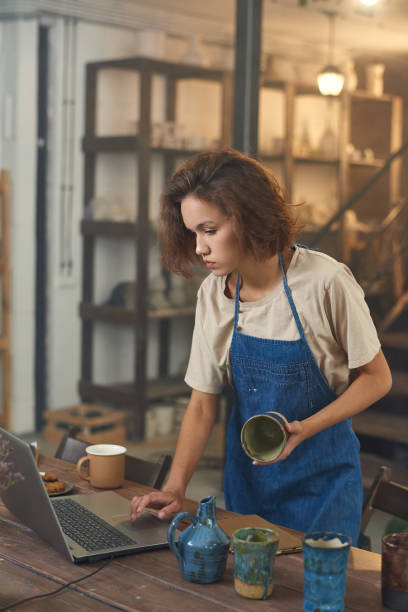 istock Craftswoman standing by table and looking at laptop screen 1353393613