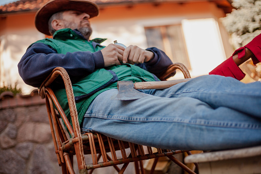 man in a hat is resting in a wicker chair in the yard in front of a pile of chopped wood. He holds a round metal flask in his hands. There is an ax on my feet