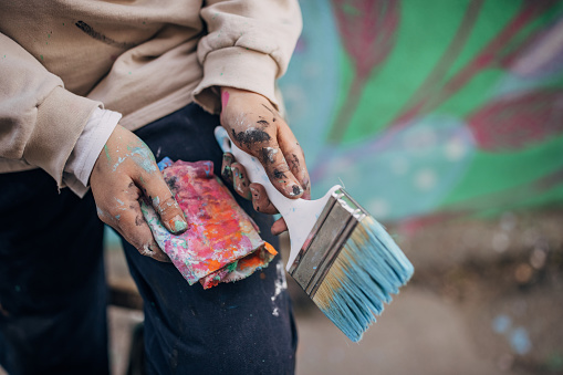 One woman, young female street artist with dirty hands holding paintbrush and rag outdoors.