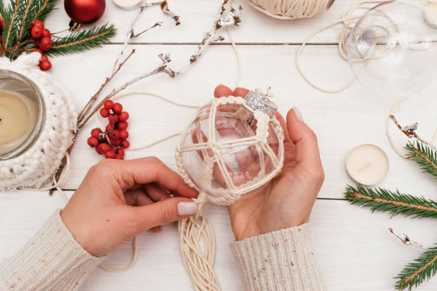 A woman decorates a transparent Christmas ball with macrame-style weaving,handmade decor in eco-style,hands close-up,top view.Christmas, New Year and eco-friendly concept stock photo