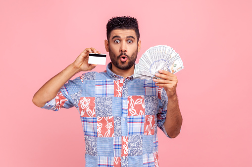 Shocked excited man with beard in blue casual shirt holding credit card and smartphone, looking at camera with big eyes, surprised with easy banking. Indoor studio shot isolated on pink background.