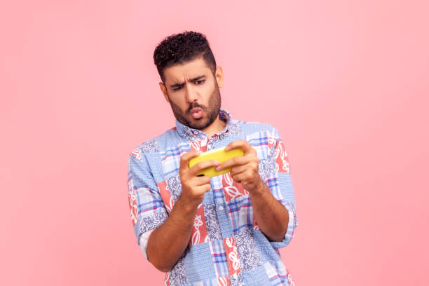 Addicted nervous bearded man wearing casual style shirt grimacing holding smart phone, playing video games online, virtual competition. Addicted nervous bearded man wearing casual style shirt grimacing holding smart phone, playing video games online, virtual competition. Indoor studio shot isolated on pink background. defeat photos stock pictures, royalty-free photos & images