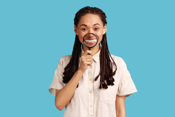 woman with dreadlocks holding magnifier on her teeth, looking at camera with happy expression. - loupe quality control magnification glass imagens e fotografias de stock