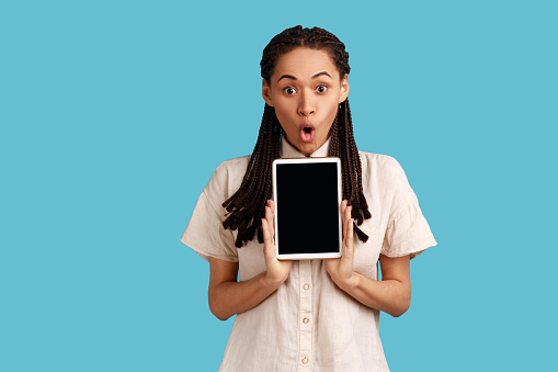 Portrait of shocked astonished woman with black dreadlocks, holding showing tablet, looking at camera with open mouth and surprise, wearing white shirt. Indoor studio shot isolated on blue background.
