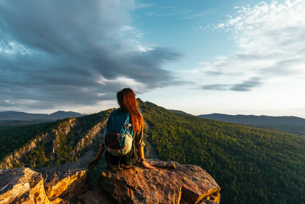 A young female tourist with a backpack admires the sunset from the top of the mountain. A traveler on the background of mountains. A traveler on the background of mountains at sunset. Copy space stock photo