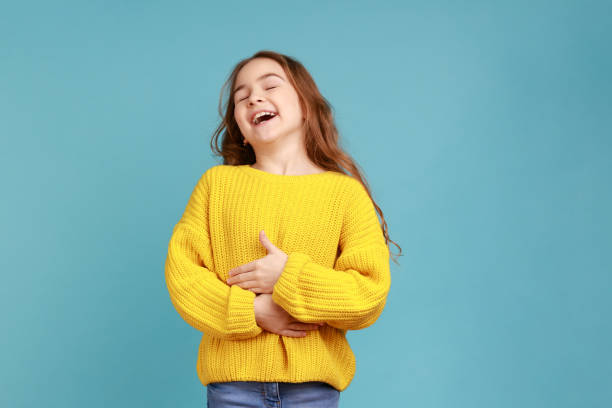 Portrait of happy positive little girl laughing out loud, holding her belly, hearing funny joke. Portrait of happy positive little girl laughing out loud, holding her belly, hearing funny joke, wearing yellow casual style sweater. Indoor studio shot isolated on blue background. child laughing hysterically stock pictures, royalty-free photos & images