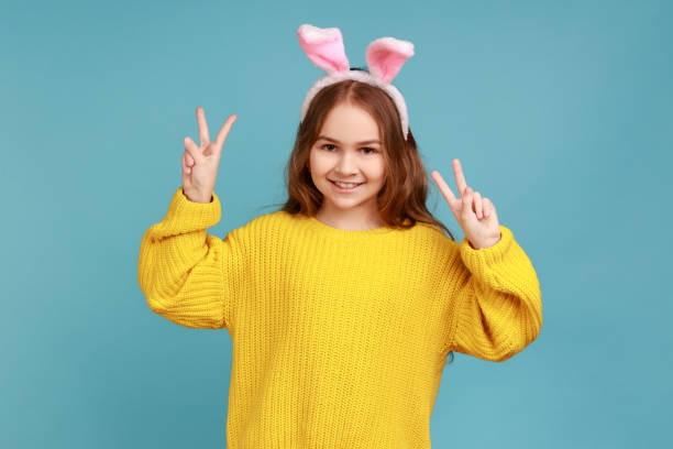 小さな女の子の肖像画は、ピンクのバニーの耳で立って、子供は楽しんで、カメラにvサインを示しています。 - easter easter bunny fun humor ストックフォトと画像