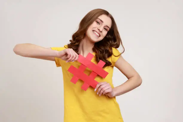 Photo of Cheerful satisfied teenager girl in yellow t-shirt showing holding red hashtag sign, being happy with target advertisement, easy blog search.