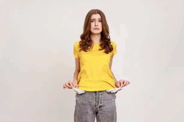 Portrait of frustrated worried brown haired female of young age in casual yellow T-shirt turning out empty pockets showing no money gesture. Indoor studio shot isolated on gray background.