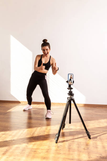 istock Fitness vlogger recording tutorials for sporty class, showing thumbs up to phone camera on tripod. 1353398993