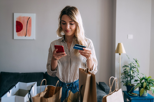 Young female making credit card purchase on her smartphone while standing with few shopping bags on table in living room