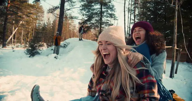 Two attractive multiethnic happy women smile sledging on snow slope towards camera, winter weekend fun slow motion. Enjoying active vacation leisure outdoors sledding down forest hill on sunny day.