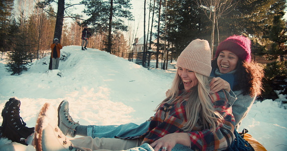 Two attractive multiethnic happy women smile sledging on snow slope towards camera, winter weekend fun slow motion. Enjoying active vacation leisure outdoors sledding down forest hill on sunny day.