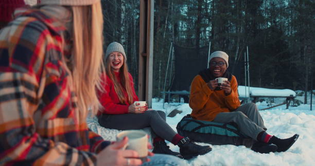 Cheerful happy multiethnic friends drink tea, talk smiling sitting on beanbags at cozy winter house terrace slow motion.