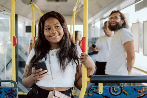 Photo of A dark-skinned woman rides in a public transport bus holding on to the railing, a girl buys an electronic ticket on her phone through an app looks at vehicle number smiles while easily making payment