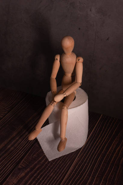 A wooden man is sitting on a roll of toilet paper in a dark room. The doll is experiencing stomach problems and intestinal diseases. A wooden man is sitting on a roll of toilet paper in a dark room. The doll is experiencing stomach problems and intestinal diseases. кукла stock pictures, royalty-free photos & images