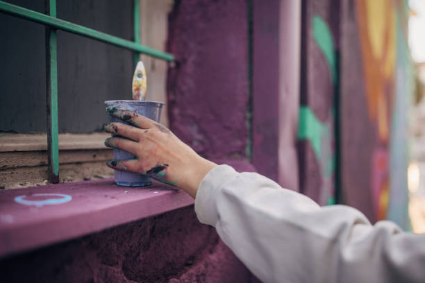 One woman, young female street artist grabbing plastic glass with paint for painting on a building wall outdoors.