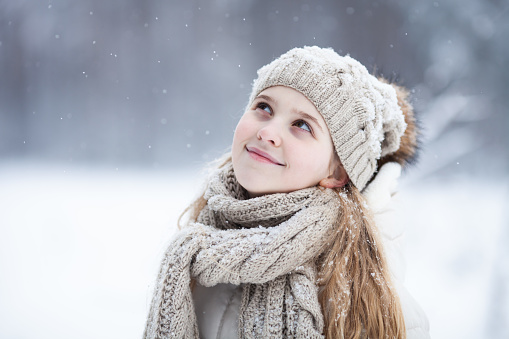 Portrait of a cute blond girl wearing warm knitted hat and scarf looking up on winter snowy background. Love winter, make a wish, dreams come true concept.