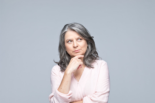 Portrait of beautiful grey hair mature woman looking up with hand on chin. Studio shot of female entrepreneur against grey background.