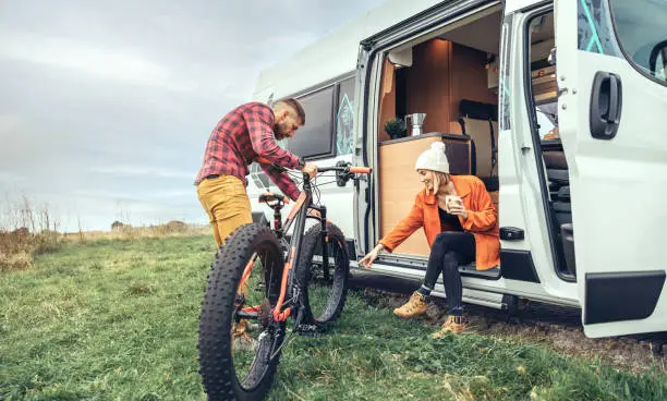 Woman drinking coffee sitting at the door of a camper van and man checking fat bike