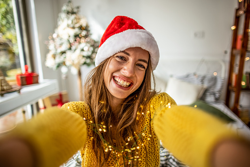 Photo of a smiling young woman with Santa hat taking selfie with mobile phone on a  Christmas morning.