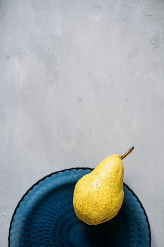 Close up one ripe yellow pear in blue glass plate on grey background. Modern minimal still life with one yellow pear. Copy space.