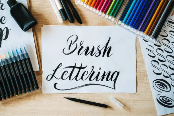 Photo of Brush Lettering text on paper and brushes and colored pencils on the table. Lettering art and calligraphy. Handwritten text Brush Lettering on white paper