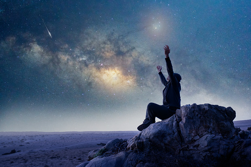 unknown person sitting on the rock with open arms or hands up under the Milky Way at night celebrating or showing amazement for the moment
