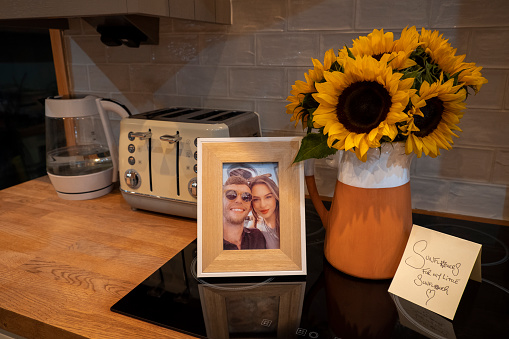 A jug filled with sunflowers, a photo of a couple in a picture frame and a cute note on a kitchen bench.