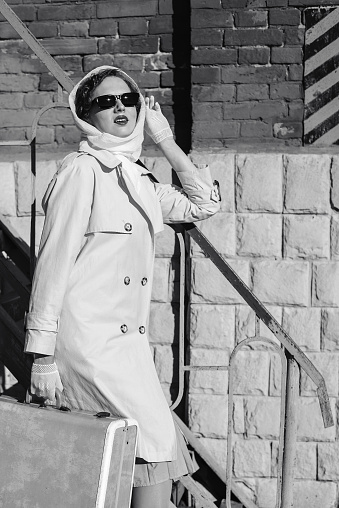 A young woman in a raincoat and dark glasses, with a suitcase in her hands, against the background of a brick wall. Retro style black and white outdoor shot