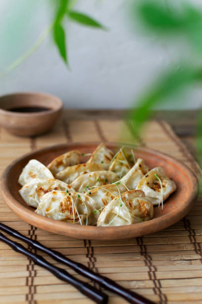 Gyoza with sesame seeds and micro greens in a ceramic bowl with bamboo sticks on a wooden table. Rustic style. Traditional Orietnal food concept. Vertical orientation. Selective focus. Gyoza with sesame seeds and micro greens in a ceramic bowl with bamboo sticks on a wooden table. Rustic style. Traditional Orietnal food concept. Vertical orientation. Selective focus. sticky sesame chicken sauces stock pictures, royalty-free photos & images