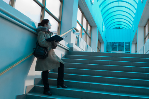 Beautiful brunette student in winter overcoat standing on a staircase inside blue light interior of some college hall under a transparent arch roof. She holds an open binder and looks at the entrance door of glass with hope and inspiration, ready to pass an exam or an interview.