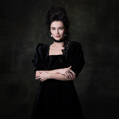 Grandeur and beauty. Portrait of young beautiful woman in image of medieval royal person in black dress isolated on dark vintage background. Concept of comparison of eras, modernity and renaissance.