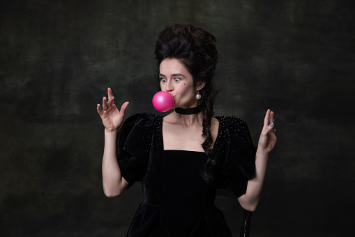 Blowing bubble gum. Classic retro portrait of young beautiful woman in image of medieval royal person isolated on dark vintage background. Concept of comparing eras, humor, modernity and renaissance.