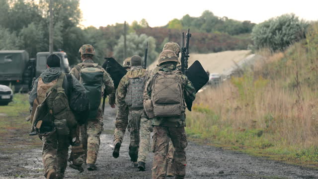 A group of military men returning after completing a task. Soldiers in military uniform with machine guns walk on a stony road with their backs to the cell