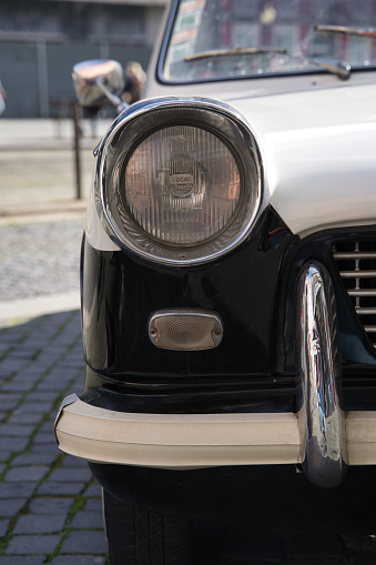 Vertical cropped photo of a headlight of a black and white classic car on a cobblestone street in a city
