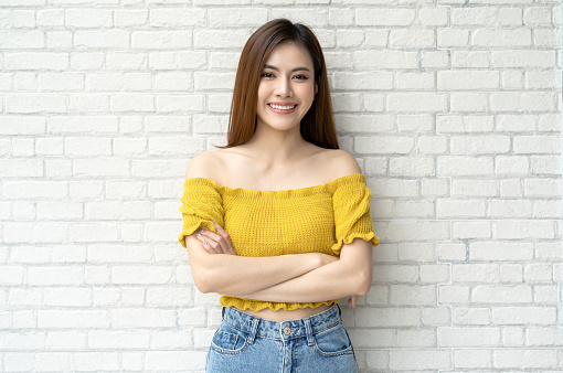 Hermosa mujer asiática sonriente vestida con ropa de punto amarilla en los hombros y posando con los brazos cruzados y mirando a la cámara sobre el fondo de la pared de ladrillo blanco. Look de invierno de moda, belleza y estilo. photo