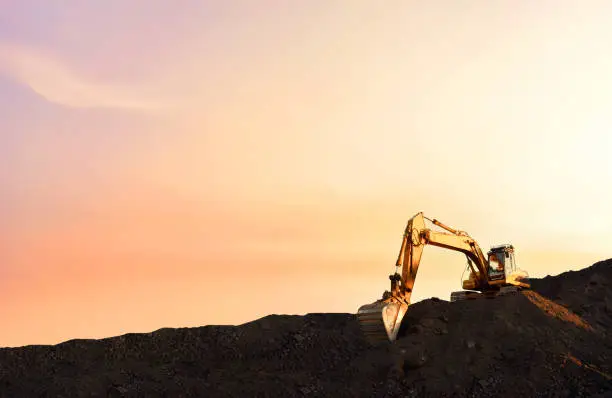 Excavator on earthmoving in open pit mining on sunset background. Backhoe digs sand and gravel in quarry. Heavy construction equipment during excavation at construction site.