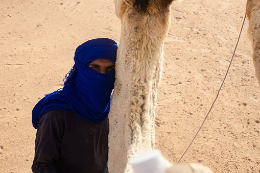 Sahara Desert, Tunisia 18,05,2021. Berber stands with a camel and looks into the distance in the Sahara Desert during a strong wind, a sandstorm.