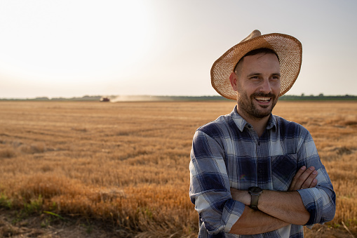 Portrait of male farmer with crossed arms wearing straw hat and standing in harvested field in summer time