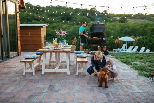 Photo of Young adult man barbecuing while his wife and daughter is playing with dog