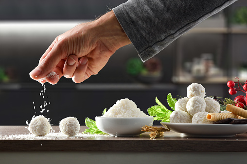 Hand pouring grated coconut over home prepared coconut balls on kitchen bench with ingredients and cooking background. Front view. Horizontal composition.