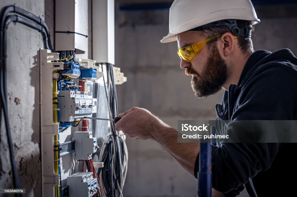 A male electrician works in a switchboard with an electrical connecting cable. A male electrician works in a switchboard with an electrical connecting cable, connects the equipment with tools. Electrician Stock Photo