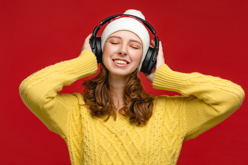 Beautiful young woman wearing Christmas Santa Claus hat is posing at the studio with red background. Person is using wireless headphones and listening New Year music. Concept with people.